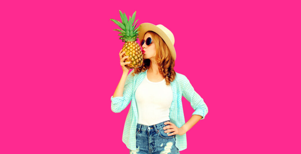 Summer portrait of woman kissing tropical pineapple wearing straw hat, sunglasses on pink background
